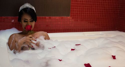 Shirtless woman smelling red rose while taking bubble bath in bathtub