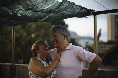Happy senior couple laughing while standing on building terrace during sunset