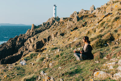 Rear view of man sitting on rock by sea against clear sky