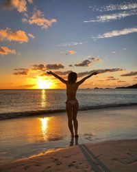 Rear view of bikini woman with arms outstretched standing at beach during sunset