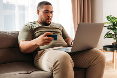 Smiling man using laptop while holding credit card sitting on sofa at home