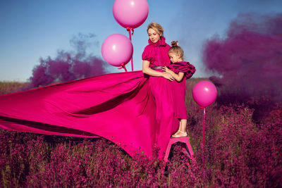 Mother and daughter in pink dresses are standing on a field with flowers at sunset  with smoke 