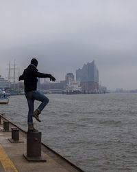 Full length of man balancing on bollard by sea at harbor against cloudy sky