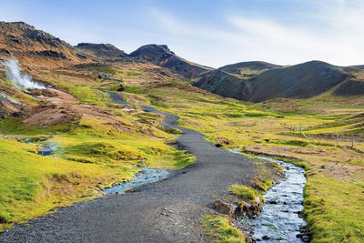 Pathway by stream amidst green landscape in volcanic valley against blue sky