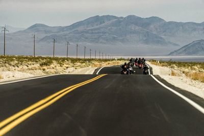 Rear view of people riding motorcycles on country road against mountains