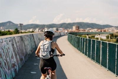 Back view confident young female bicyclist in sportswear and helmet riding bike on fenced curvy paved track while training alone in summer day