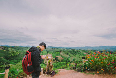 Side view of young man with backpack using mobile phone while standing on mountain against cloudy sky