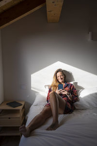Blond lady sitting on bed and browsing smartphone while resting in attic