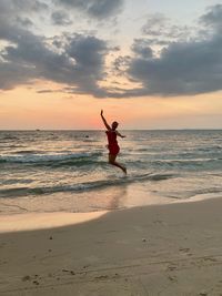 Full length of woman jumping on shore at beach against sky during sunset