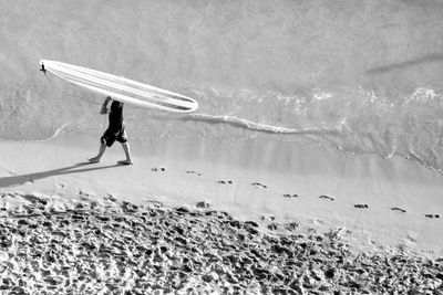 High angle view of man holding surfboard while walking on sand at beach