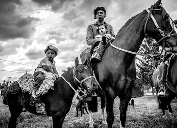 Low angle view of father and children sitting on horses against cloudy sky 