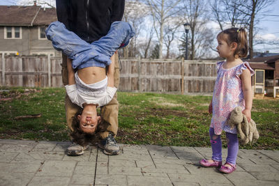 Cute girl looking at father carrying brother upside down while standing in backyard