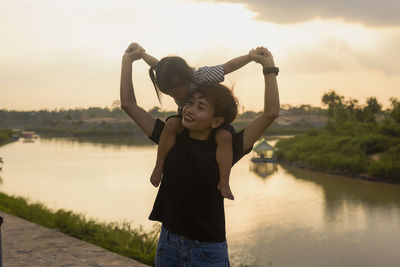 Portrait of young woman with arms raised standing against lake during sunset