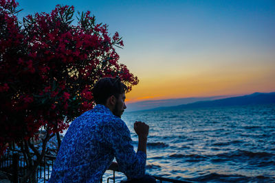 Woman sitting by sea against sky during sunset