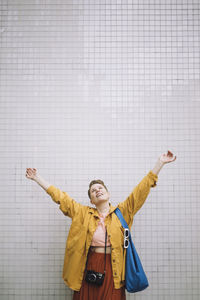 Happy mature woman with arms raised standing against wall