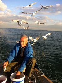 Portrait of fisherman sitting in boat while sea gulls flying against sky