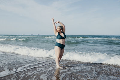 Content curvy female in bikini standing looking away on seashore and enjoying summer holiday with raised arms