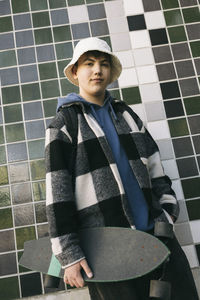 Portrait of teenage boy with skateboard standing against tiled wall