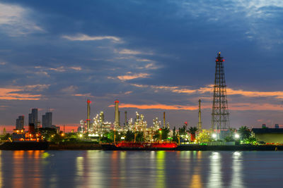 Illuminated factory by river against sky in city