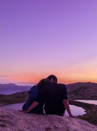 Rear view of couple romancing on rock by mountains against sky during sunset