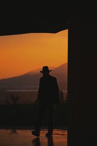 Rear view of silhouette man standing by mountain against orange sky