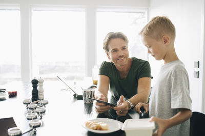 Smiling man showing digital tablet to boy at dining table against window