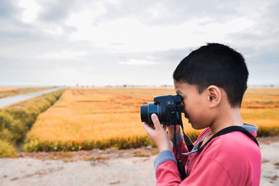 Boy photographing with camera on field against sky