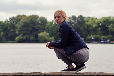 Full length portrait of beautiful woman crouching on retaining wall by river against sky