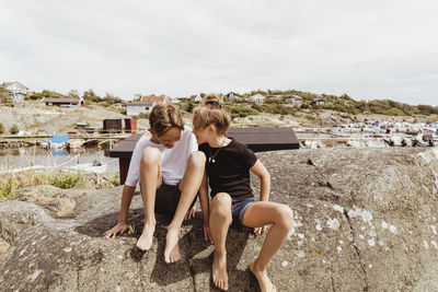 Happy siblings sitting on rock against sky during summer vacation