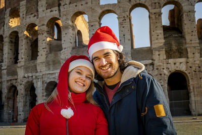 A lovely young couple taking a picture in front of the colosseum with red santa hats. 