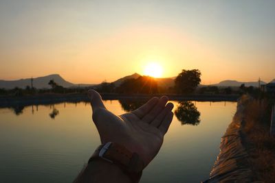 Reflection of hand in lake against sky during sunset