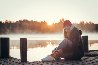 Brunette with realistic smile sits on a wooden bridge by a lake at sunrise.  paltamo, finland.