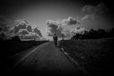 Rear view of man riding bicycle on road amidst land against cloudy sky