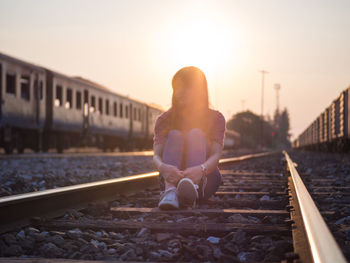 Woman sitting on railroad track against sky during sunset