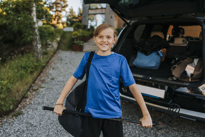 Portrait of smiling boy holding ice hockey stick while standing in front of car trunk