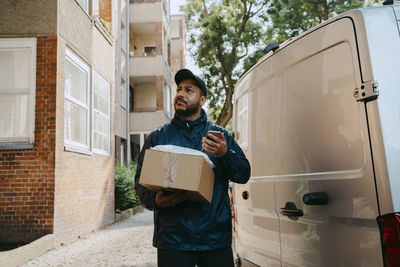 Confused male courier delivery person holding package while standing next to van near building