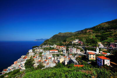 High angle view of townscape on green mountain by sea against clear blue sky