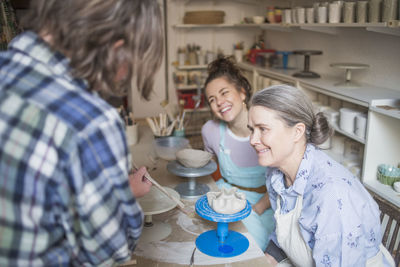 Happy female potters looking at mature colleague using hand tool at ceramic workshop
