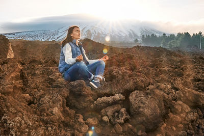 Woman enjoying freedom while meditating on lava stone at panoramic view of snowy summits of etna