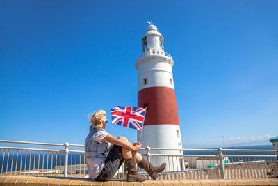 Woman holding flag while sitting by railing against sky