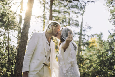Bride showing ring finger while kissing groom on mouth in forest during wedding
