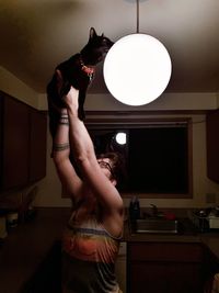 Woman holding cat by illuminated lighting equipment in kitchen at home