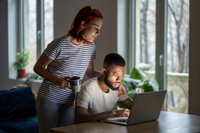 Loving woman wife standing behind husband working on laptop, supporting him in online business