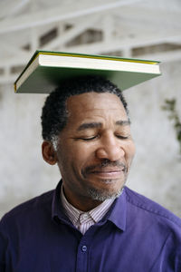 Smiling mature man with eyes closed balancing book on head