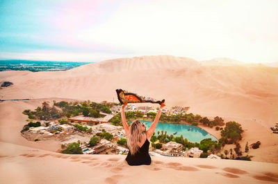 Rear view of woman sitting on sand dune against sky