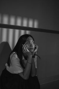 Young woman with hands on chin sitting against wall at home