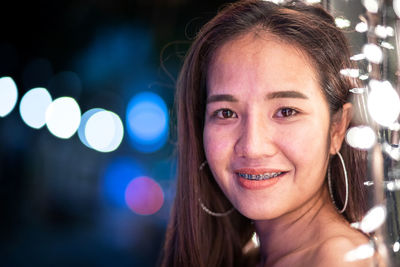 Close-up portrait of smiling young woman by illuminated string lights at night