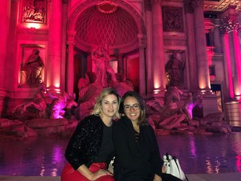 Portrait of friends sitting against fountain in illuminated city