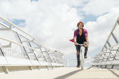 Full length of man cycling on bridge against cloudy sky in city