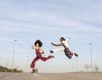 Side view of african american couple in trendy traditional clothes jumping high above asphalt road and performing dance on background of blue sky